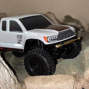 May include: A white toy truck with black wheels and a gold-colored bumper navigates a rocky terrain. The truck has the words "BASE CAMP" and other text on the side. The hood features a black and orange design.