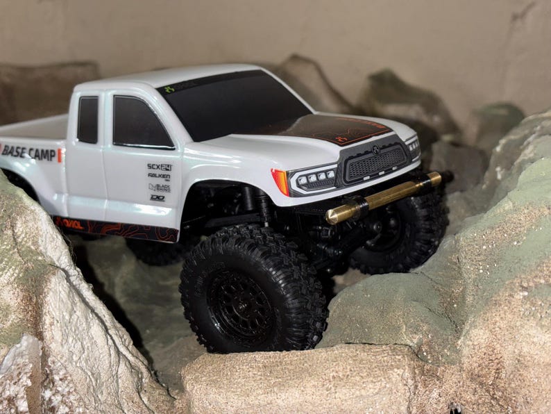 May include: A white toy truck with black wheels and a gold-colored bumper navigates a rocky terrain. The truck has the words "BASE CAMP" and other text on the side. The hood features a black and orange design.