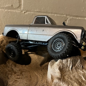 May include: A silver toy truck with black tires and a black grill. The truck is on a dirt and rock surface.