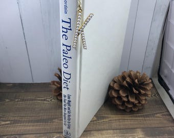 May include: A gold-colored metal bookmark with a chain and a letter "K" embellished with small, white, sparkling stones. The bookmark is placed in a book titled "The Paleo Diet" on a wooden surface.