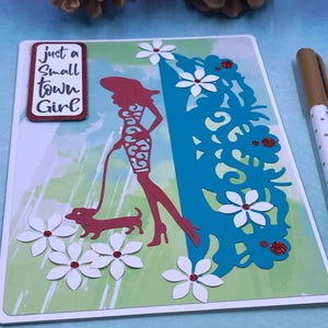 May include: A handmade greeting card featuring a red silhouette of a woman walking a dachshund. The card has white paper flowers with red centers, a blue floral cutout, and the text "just a small town girl".
