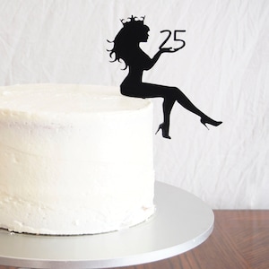 May include: White cake with pink flowers and a black silhouette cake topper of a woman wearing a crown and holding the number 25.