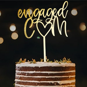 Peut inclure: Topper de gâteau en paillettes dorées avec les mots "engaged" et "&" avec un cœur au milieu. Le topper est sur un gâteau à étages avec un glaçage blanc.
