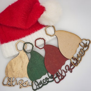 May include: Three wooden Christmas ornaments shaped like Santa hats, each painted a different color: gold, green, and red. The ornaments have names written on them: "Eliza", "Jacob", and "Ellie".