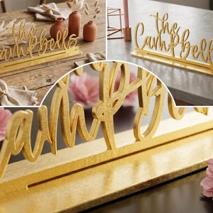 May include: A gold-colored wooden sign that reads "the Campbells" in a cursive font. The sign is displayed on a table, with a slot to hold place cards. The sign is a decorative item.