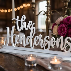 May include: A silver wooden sign that reads "The Hendersons" in cursive script. The sign is on a wooden table, with candles and a bouquet of roses in the background. The sign is a wedding or event decoration.