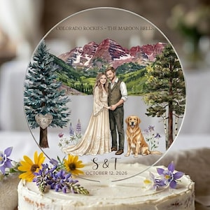 May include: A clear, circular wedding cake topper featuring a couple, a dog, and a mountain landscape. The text "COLORADO ROCKIES - THE MAROON BELLS" and the couple's initials and wedding date are displayed. The cake is decorated with flowers.