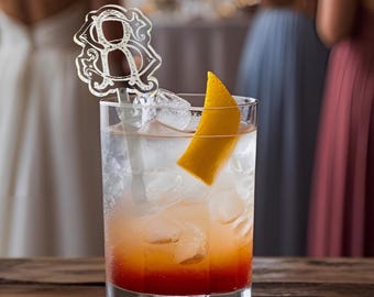 May include: A cocktail in a clear glass with ice, an orange peel garnish, and a decorative monogrammed stirrer. The drink has a layered appearance with red, orange, and clear liquids. The glass sits on a wooden surface.