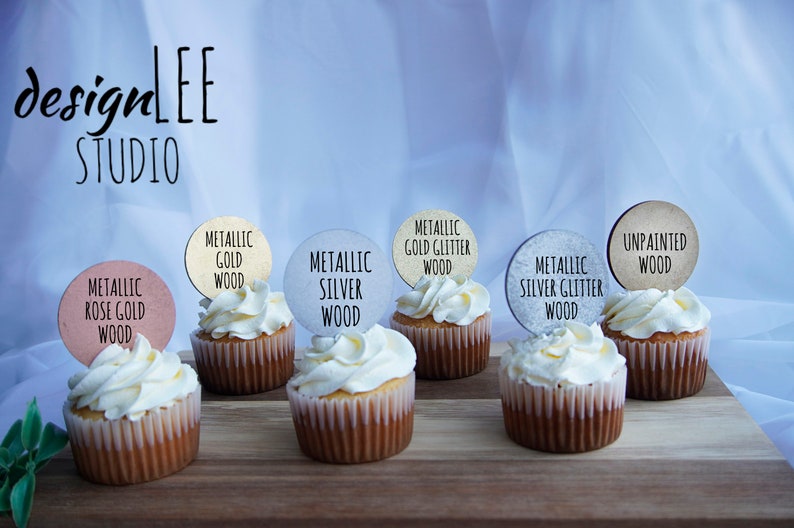 May include: Cupcakes with white frosting and wooden toppers labeled "Metallic Rose Gold Wood", "Metallic Gold Wood", "Metallic Silver Wood", "Metallic Gold Glitter Wood", "Metallic Silver Glitter Wood", and "Unpainted Wood". The image includes the "designLEE STUDIO" logo.