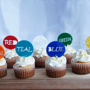 May include: Cupcakes with white frosting and colorful toppers. The toppers are round and feature the colors orange, red, teal, white, blue, green, clear, and yellow, with the color names printed on them.