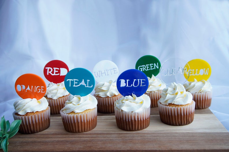 May include: Cupcakes with white frosting and colorful toppers. The toppers are round and feature the colors orange, red, teal, white, blue, green, clear, and yellow, with the color names printed on them.