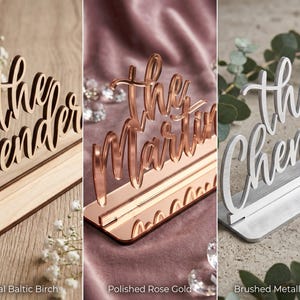 May include: Three wedding table signs in different styles. One is natural Baltic birch, another is polished rose gold, and the third is brushed metallic silver. Each sign displays a family name in elegant script.