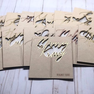 May include: A collection of personalized cake toppers with names and hearts cut out of wood. The toppers are on a brown background and are ready to be used for a wedding or special occasion.