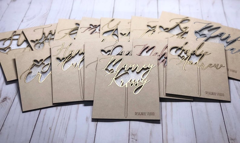 May include: A collection of personalized cake toppers with names and hearts cut out of wood. The toppers are on a brown background and are ready to be used for a wedding or special occasion.