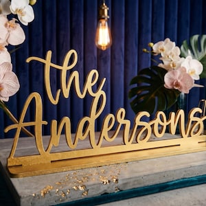 May include: A gold-colored wooden sign that reads "the Andersons" in a cursive font. The sign is on a gray surface with gold leaf accents, with flowers and candles in the background.