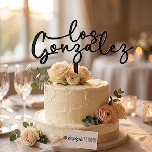 May include: A two-tiered white frosted cake decorated with fresh roses and greenery. A black cake topper reads "Los Gonzalez" in cursive script. A tag with "designdee STUDIO" is attached to the cake.