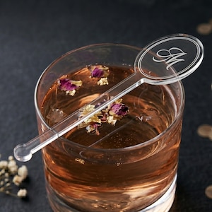 May include: A clear acrylic cocktail stirrer with an oval top engraved with the letter "M" rests in a glass of amber-colored liquid. The drink contains rose petals and gold leaf flakes, creating an elegant presentation.