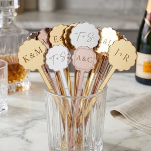 May include: A collection of personalized drink stirrers in a clear glass. The stirrers have scalloped tops in gold, silver, and rose gold, each engraved with initials. The initials include "K&M", "E&S", "T+S", "A&C", and "J+R".