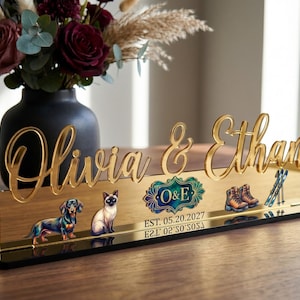 May include: A gold-colored acrylic desk sign with the names "Olivia & Ethan" in elegant script. The sign features illustrations of a dachshund, a Siamese cat, hiking boots, and skis. The sign also includes the text "O&E" and the date "05.20.2027".