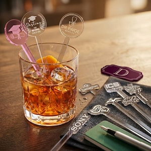 May include: A glass of amber-colored liquid with ice and an orange slice, with several clear acrylic drink stirrers. The stirrers have various designs and text, including a graduation cap, the word "Destiny", and the name "Dr. Paige".