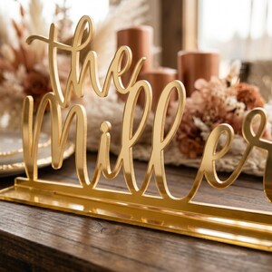 May include: A gold-colored table sign that reads "The Millers" in elegant cursive script. The sign is placed on a wooden table, with blurred background elements including candles and floral arrangements.