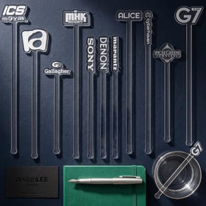 May include: A collection of clear acrylic drink stirrers with various logos and text, including "ICS", "MHK WEEKENDS", "ALICE", "SONY", "DENON", "marantz", "Gallagher", "Cyberhaven", "Spicer Greene Jewelers", and "G7". Also includes a green notebook, a silver pen, and a black business card.