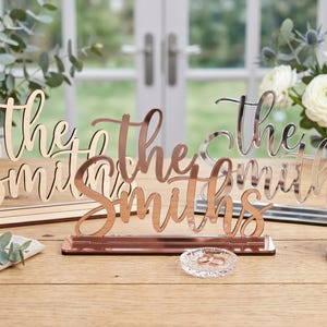 May include: Three decorative signs that read "The Smiths" in a cursive font. One is made of wood, one is rose gold, and one is silver. The signs are displayed on a wooden table, with a small glass dish and flowers in the background.