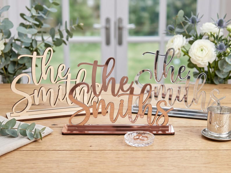 May include: Three decorative signs that read "The Smiths" in a cursive font. One is made of wood, one is rose gold, and one is silver. The signs are displayed on a wooden table, with a small glass dish and flowers in the background.