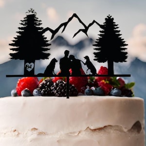 May include: Black silhouette cake topper featuring a couple, two dogs, a mountain range, and two pine trees. The couple is holding hands and the dogs are sitting next to them. The topper is on a white cake with berries and blueberries.