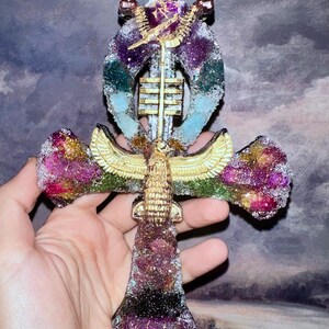 Könnte beinhalten: Ein Ankh-Symbol-Ornament mit einer texturierten, mehrfarbigen Glitzer-Oberfläche in Lila, Pink, Grün und Blau. Die Mitte ist mit einem goldfarbenen geflügelten Skarabäus und einer vertikalen Säule aus horizontalen Goldbalken verziert.