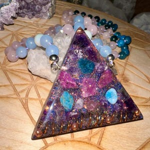 Op de afbeelding: Een driehoekige kristallen hanger met een koperen draadwikkeling en een verscheidenheid aan paarse, roze en blauwe kristallen ingebed in helder hars. De hanger hangt aan een kralenketting met roze, blauwe en groene kralen.