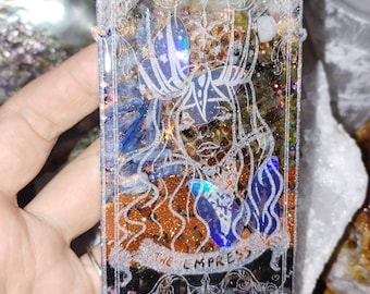 May include: A rectangular resin tarot card featuring the Empress. The card has a silver border with intricate designs. The interior has a mix of colors, including blue, orange, and gold glitter. The words "THE EMPRESS" are at the bottom.