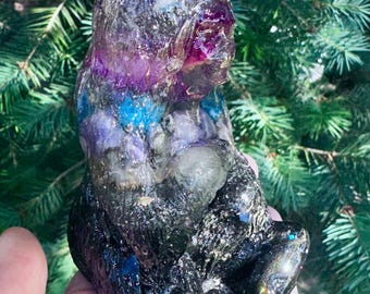 May include: A translucent resin figurine shaped like a seated bear, featuring a swirling mix of deep purple, bright blue, and black colors with embedded gold flakes and glitter throughout the textured, glossy surface of the sculpture.
