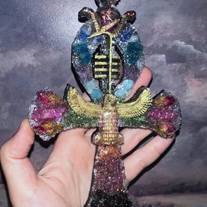 May include: An ankh-shaped decorative object featuring a textured, glittery surface in shades of purple, pink, and black. It includes a central gold-colored winged figure, a vertical gold-colored bar, and embedded blue and purple crystal-like accents.