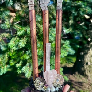 May include: A cylindrical resin base filled with purple stones and gold flakes, supporting three copper pipes wrapped in copper wire and topped with clear quartz points. A central selenite wand and a small rose quartz angel figurine are included.