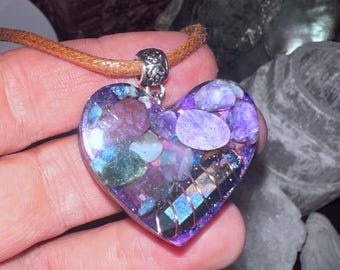 May include: A heart-shaped pendant necklace with a brown leather cord. The pendant is filled with purple and blue stones and resin, with a copper wire design around the edges. The pendant is approximately 2 inches wide.