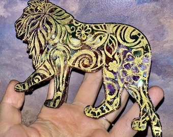 May include: A decorative lion figurine with intricate gold filigree and floral patterns. The lion is in a walking pose, with a mix of purple, green, and gold glitter filling the open spaces. The figurine is held in a hand.