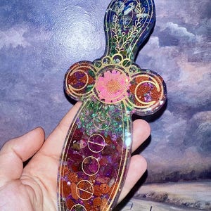 May include: A decorative dagger-shaped resin pendant with a celestial theme. The pendant features a handle with gold filigree and a blade filled with colorful stones and glitter. The design includes a pink star, crescent moons, and other celestial symbols.