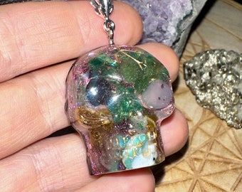 May include: A clear resin skull pendant with a silver bail, strung on a black cord. The skull is filled with green, gold, and purple inclusions. The pendant is displayed against a background of amethyst and pyrite.