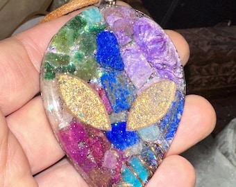 May include: A resin pendant shaped like an alien head, with a variety of colourful gemstones embedded in the resin. The pendant is on a brown leather cord and has a silver bail.