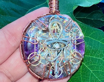 May include: A circular pendant featuring a silver-toned ankh and Eye of Horus symbol embedded in a resin base with purple and clear crystalline elements, gold-colored wire geometric patterns, and copper wire wrapping with a matching cord.