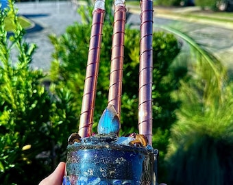May include: A cylindrical resin base filled with layers of blue, gold, and pink materials, topped with a blue iridescent stone and three copper-wrapped rods, each capped with a clear, pointed crystal at the top of the structure.