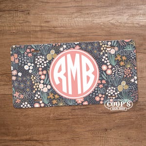May include: A license plate with a floral design in navy, pink, and yellow. The center features a pink circle with the white letters "RMB". The bottom right corner has the text "COOP'S SIGN SHOP".