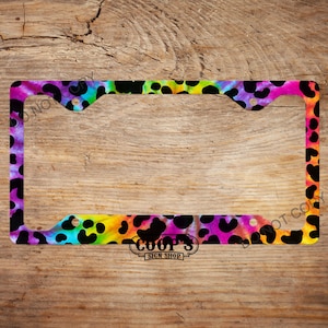 May include: License plate frame with a vibrant tie-dye and leopard print design. The frame features a rainbow of colors, including pink, purple, yellow, and green, with black leopard spots. The text "COOP'S SIGN SHOP" is visible.