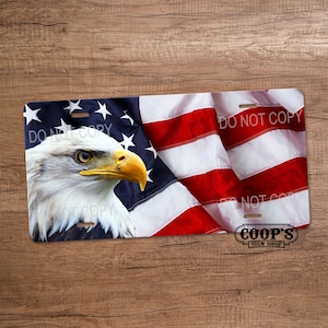 May include: A metal license plate featuring a bald eagle with a close-up view of its head and beak. The eagle is positioned in front of a waving American flag with red and white stripes and blue stars. The text "DO NOT COPY" appears on the plate. The bottom right corner of the plate has the text "COOP'S SIGN SHOP".