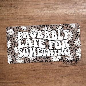 May include: Rectangular license plate with a brown leopard print background and white daisy flowers. The text "PROBABLY LATE FOR SOMETHING" is in large white letters. The sign also has the text "COOP'S SIGN SHOP".