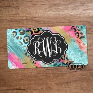 May include: A license plate with a colorful design featuring a black and white monogram. The background has a mix of teal, pink, and gold with leopard print accents. The plate also has the text "COOP'S SIGN SHOP".