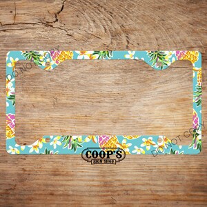 May include: License plate frame with a tropical design. The frame has a light blue background with a pattern of yellow pineapples, white flowers, and green leaves. The text "COOP'S SIGN SHOP" is at the bottom.