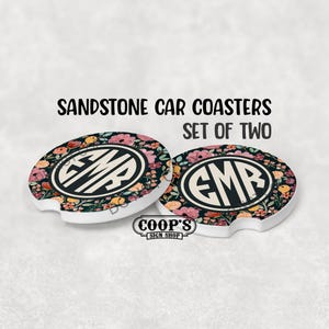May include: Set of two sandstone car coasters. Each coaster features a floral design with pink, peach, and yellow flowers on a dark background. The center has a black and white monogram. The text "SANDSTONE CAR COASTERS SET OF TWO" is above the coasters.