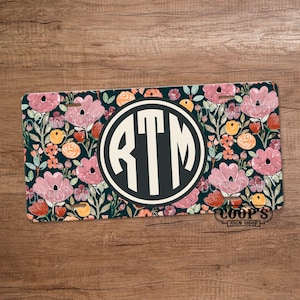 May include: Rectangular license plate with a floral design. The background is a dark teal with pink flowers, orange and yellow accents, and green leaves. A white circle in the center contains the letters "RTM" in a dark teal font.